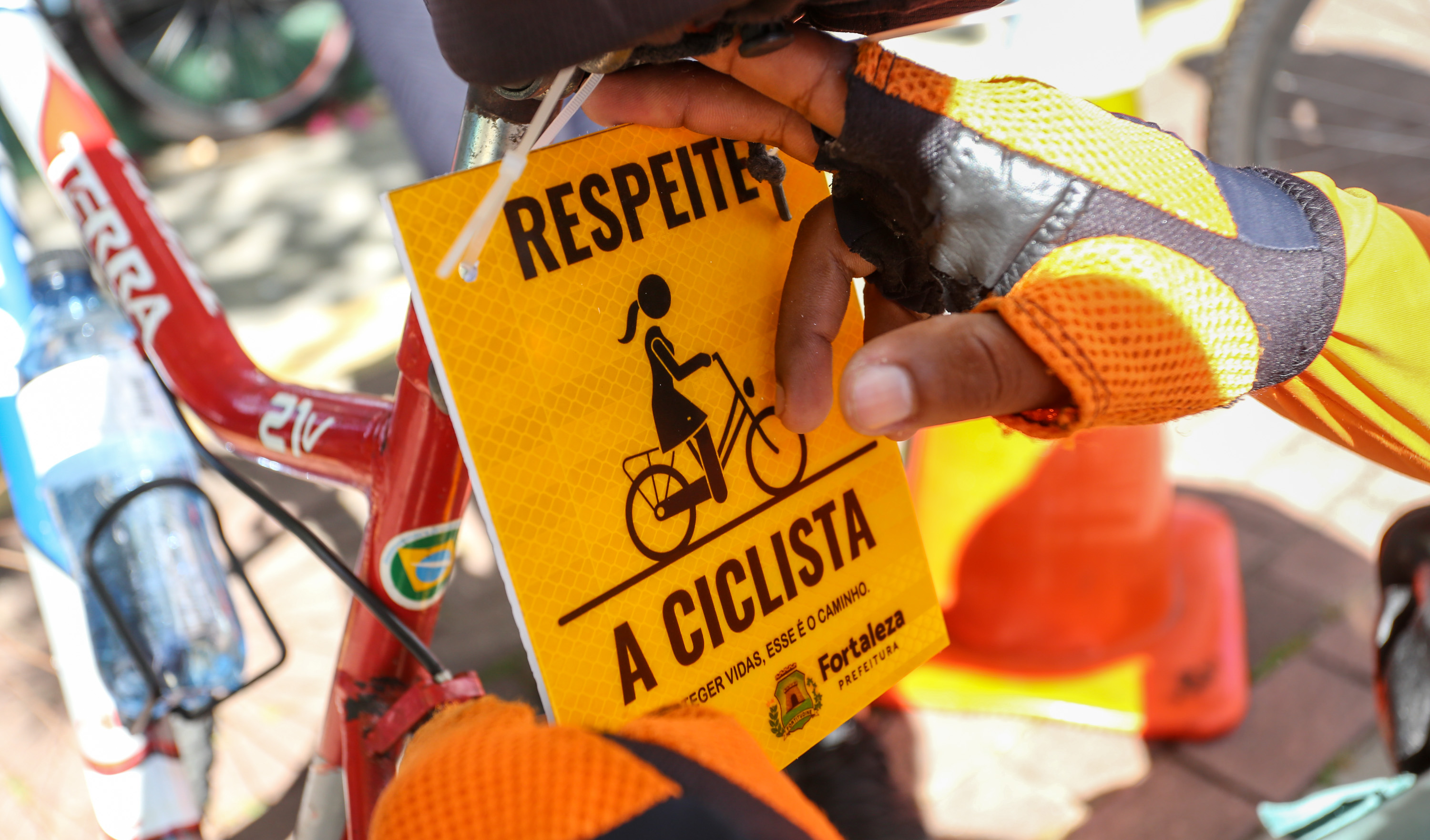 a foto mostra duas mãos colocando uma placa amarela com a frase respeite a ciclista atrás do selim de uma bicicleta